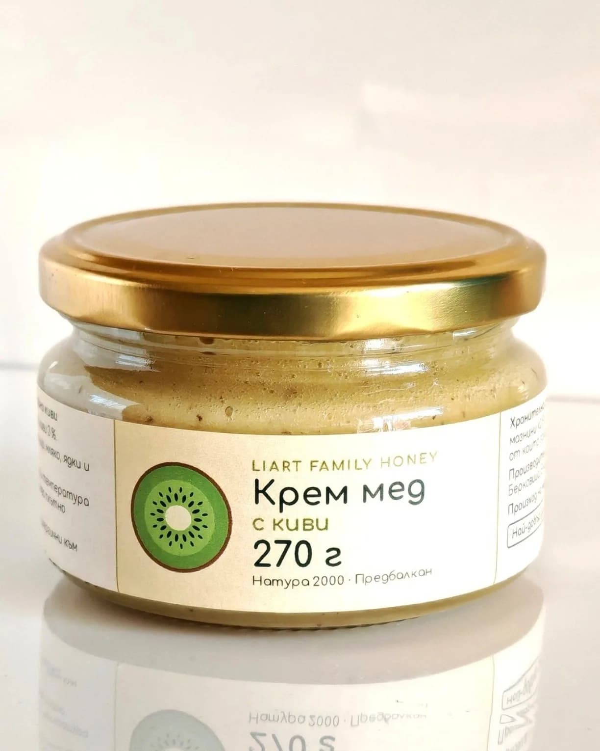 Стъклен буркан мед със златистa капaчка и жълтеникав кремообразен мед със ситни тъмни точици, на бял фон.